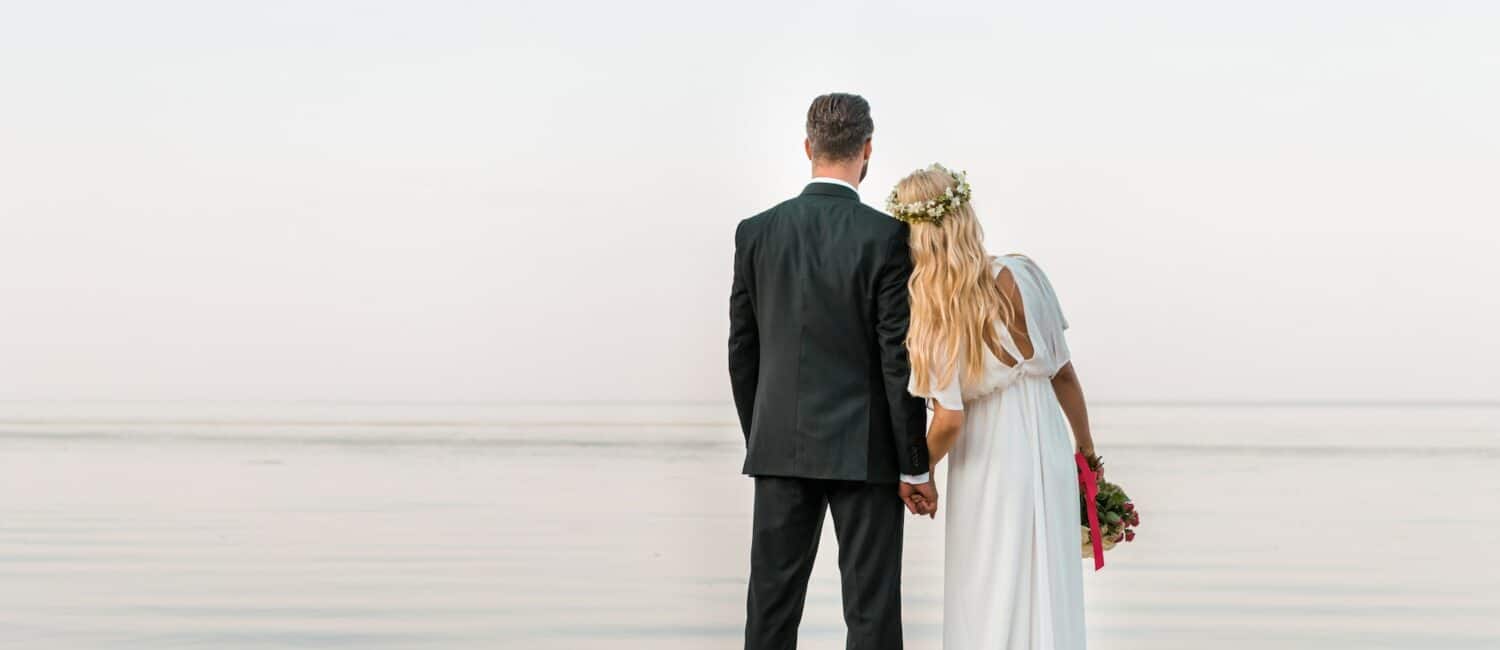 back view of wedding couple standing on beach with wedding bouquet and looking at sea